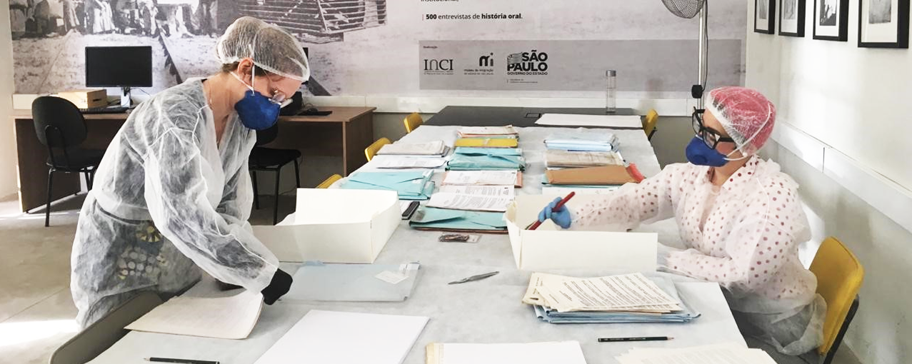 Colaboradoras do Museu estão mexendo em documentos do arquivo, utilizando capas, máscaras e luvas
