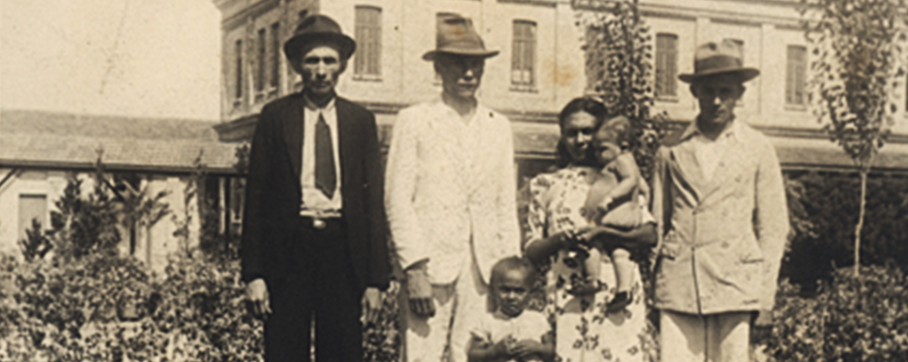Fotografia de acervo, em sépia, mostrando uma família formada por três homens, uma mulher e duas crianças, posando com a Hospedaria do Brás e um jardim de fundo