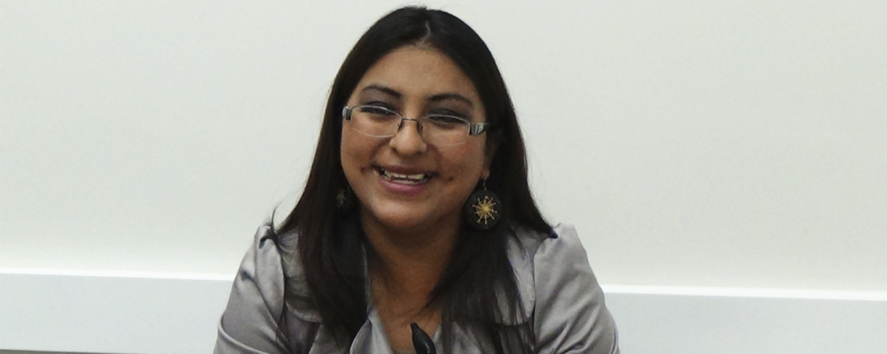 A fotografia mostra uma mulher, de origem boliviana, com cabelos escuros um pouco abaixo do ombro, blusa cinza e óculos. Ela está sorrindo