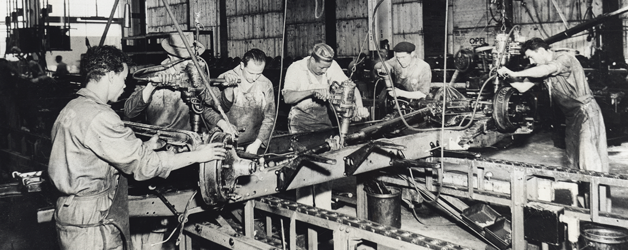 Em uma fábrica, seis homens operam maquinários em um dia de trabalho