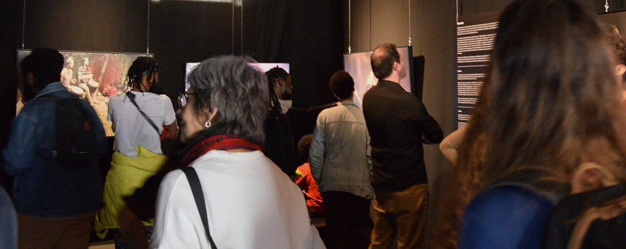 Na fotografia tirada durante a inauguração, diversos visitantes contemplam as imagens apresentadas na exposição "Pertencimentos Transnacionais: Movimentos e Ritmos na Música Africana".