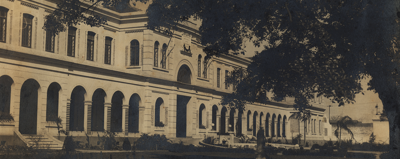 Imagem retangular, em sépia, mostrando, de lado, a fachada do edifício da antiga Hospedaria de Imigrantes do Brás, com sombra de árvore à direita