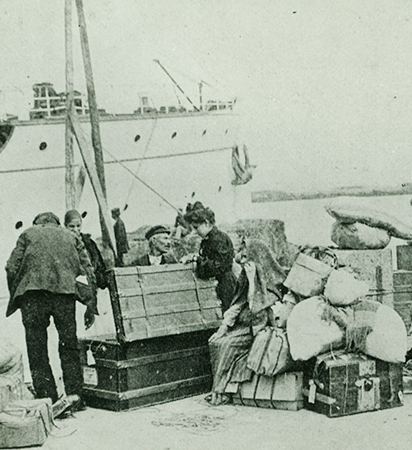 Fotografia de acervo, em preto e branco, mostrando um grupo de migrantes italianos, na margem de um porto, em volta de baús e malas, com parte de um navio ao fundo e à esquerda