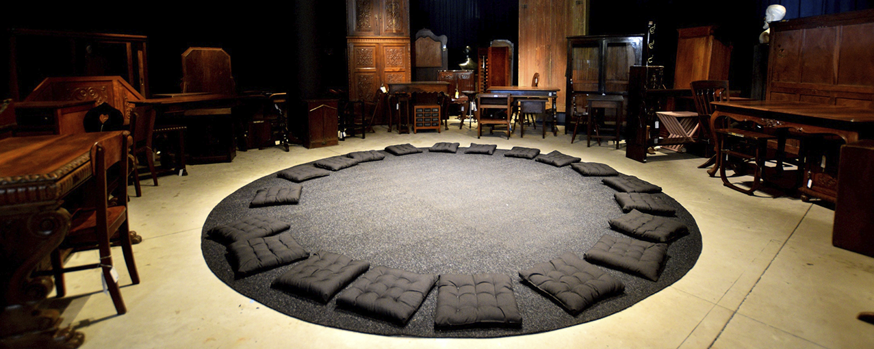 Móveis de madeira estão dispostos em um semicírculo com um tapete redondo cinza escuro no chão e almofadas da mesmas cor em cima dele