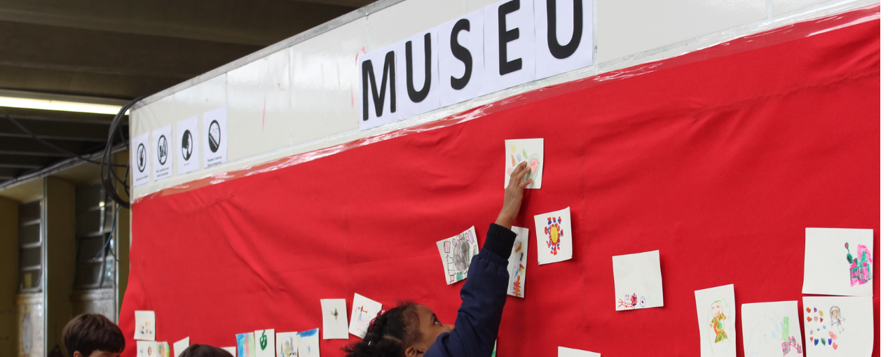 Na imagem consta alunos dispostos à frente de um mural, realizando uma atividade educativa.