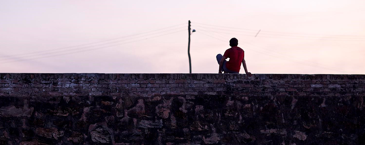 A imagem mostra, de costas, um menino sentado em cima de um muro. Na frente dele, é possível ver um poste com fios elétricos