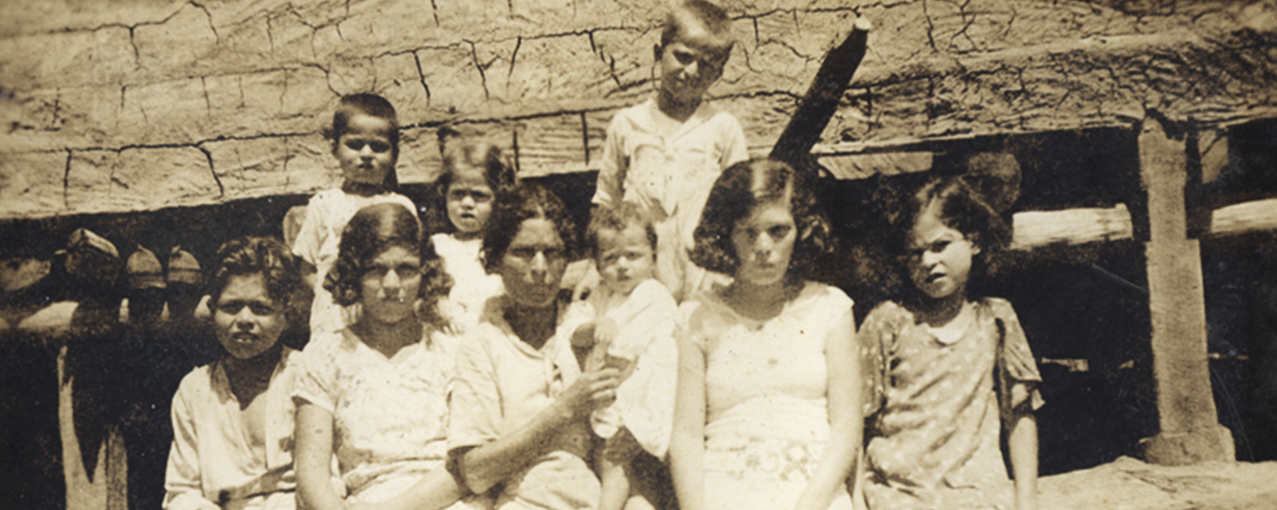 Fotografia retangular, em sépia, mostrando três crianças atrás e cinco crianças e uma mulher à frente. Ao fundo, uma estrutura retangular horizontal de concreto, com várias rachaduras