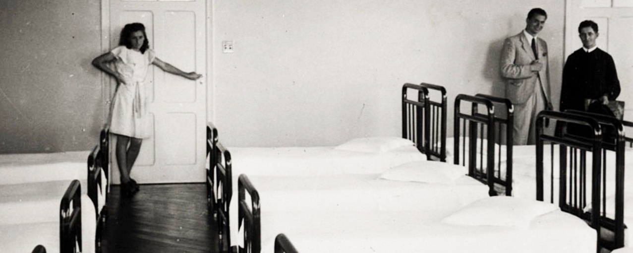 A imagem, em preto e branco, mostra camas em uma enfermaria. Uma enfermeira e dois homens estão em pé no fundo da fotografia