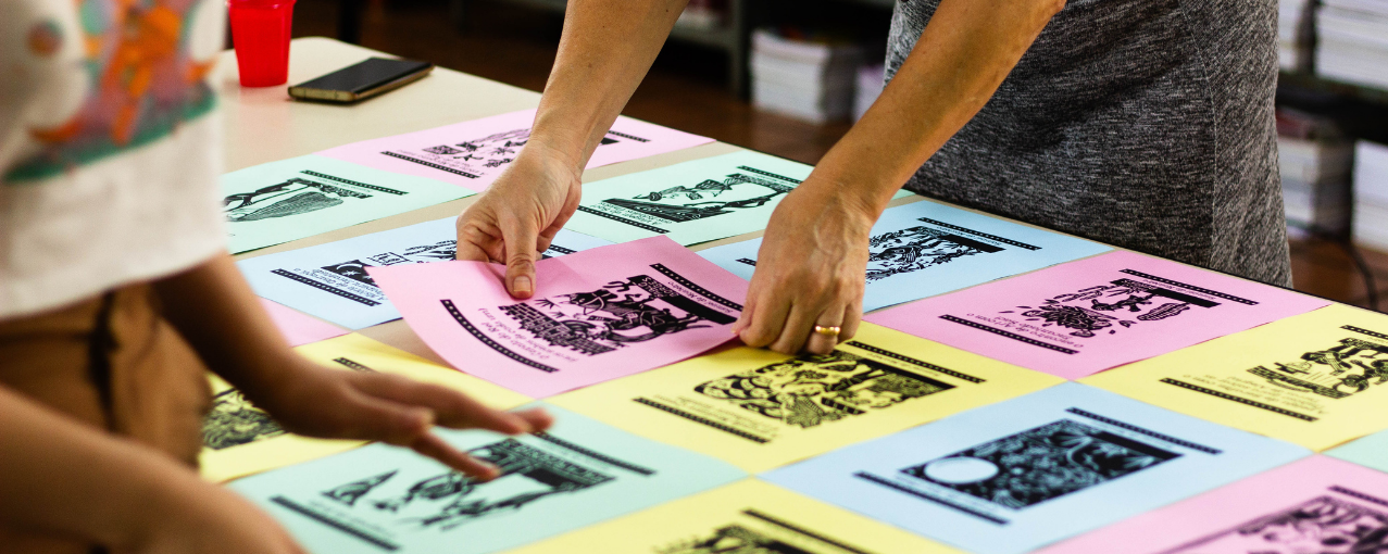 Mãos organizando folhetos de cordel coloridos, com capas ilustradas em xilogravura, sobre uma mesa